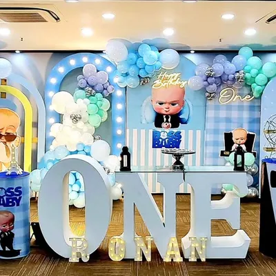 A grand Boss Baby first birthday party setup. The design includes a full stage with multiple character cutouts, a large light-up 'ONE', and coordinated balloon arches in a pastel blue and mint palette.