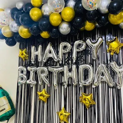 A classic and festive "Happy Birthday" setup perfect for any home party. This decoration features a black fringe curtain, a silver foil "Happy Birthday" banner, and a ceiling garland of black, gold, and silver balloons.