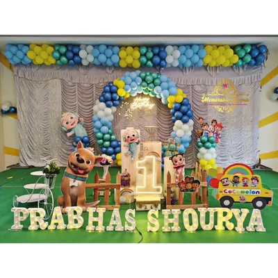 A grand Cocomelon theme for a first birthday party, complete with character cutouts, a custom backdrop, and a balloon arch in the show's signature colors. The setup includes a light-up number '1' and the birthday boy's name in marquee letters.