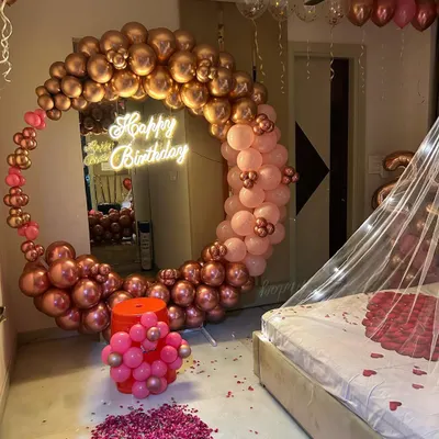 A romantic birthday room decoration featuring a large balloon ring in chrome rose gold. The setup includes a 'Happy Birthday' neon sign and a path of rose petals, creating a perfect surprise.