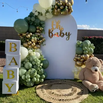 A beautiful outdoor baby shower setup with a modern, rustic feel. I used a balloon arch in sage green, white, and gold, along with "BABY" blocks and a large teddy bear.