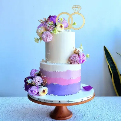 A two-tiered wedding or engagement cake with a purple ombre effect. This design is decorated with artificial flowers and a gold ring topper, but I teach how to create similar flowers from sugar.