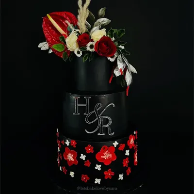 A black wedding cake is a statement. This dramatic three-tiered creation features a cascade of red and white sugar flowers and a custom monogram, proving that wedding cakes can be both bold and beautiful.