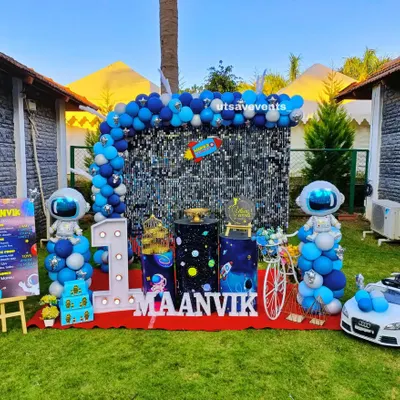 An exciting outdoor space-themed first birthday party. The setup features a large sequin backdrop, balloon astronaut figures, a custom milestone board, and even a miniature car for the birthday boy.