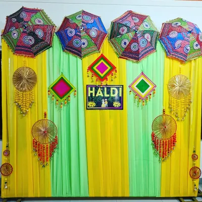 A colorful and traditional Haldi backdrop. This setup features yellow and green drapes, decorative umbrellas, and handcrafted hangings for a festive look.