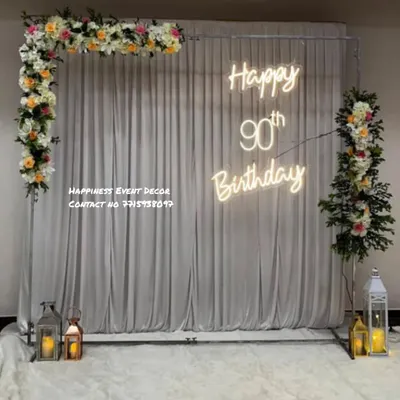 An elegant and classy setup for a 90th birthday celebration. This decor features a simple frame with a white curtain backdrop, floral arrangements, lanterns, and a custom neon sign.