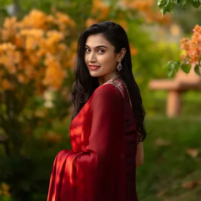 An elegant portrait of a woman in a deep red saree, looking over her shoulder with a gentle smile against a backdrop of blooming orange flowers.