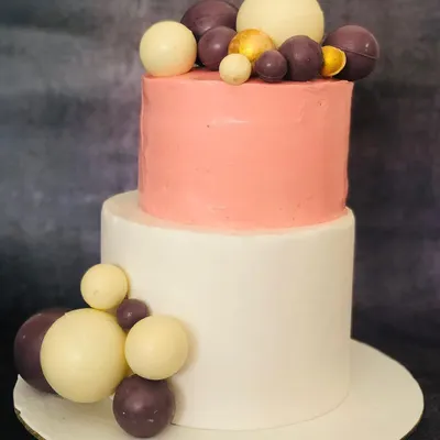 This two-tiered cake combines a soft pink top tier with a clean white bottom tier. The cascading chocolate spheres in white, purple, and gold add a modern, playful touch to an elegant design.