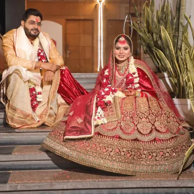 A regal portrait of the bride and groom on their wedding day. The bride is seated on the steps, allowing her magnificent red and gold lehenga to be displayed in its entirety, while the groom sits beside her in his traditional attire. This posed shot captures the grandeur of the occasion.