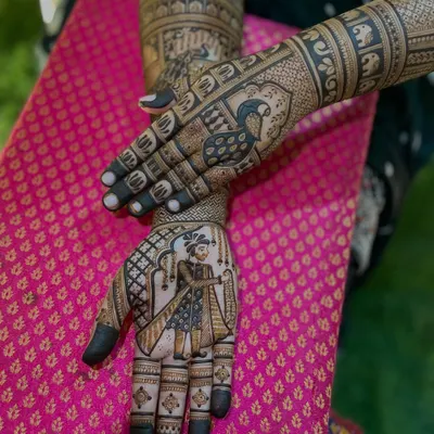 A truly personal bridal mehendi featuring a portrait of the groom on one hand and a peacock on the other. The arms are adorned with elephant motifs and jharokha patterns, creating a royal narrative.