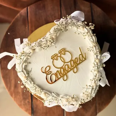 An elegant heart-shaped engagement cake. It's adorned with delicate baby's breath, white ribbons, and a golden "Engaged" topper for a classic, romantic look.