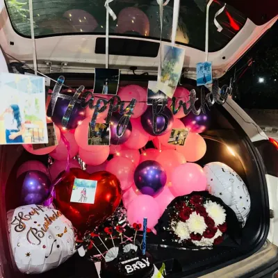 A car trunk filled to the brim for a birthday surprise, including pink and purple balloons, a 'Happy Birthday' banner, hanging photos, a cake, and a bouquet.