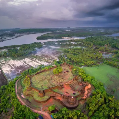 An aerial panorama of Mirjan Fort near Kumta, captured under a dramatic monsoon sky. The drone's perspective reveals the fort's strategic location, nestled amidst lush greenery and the winding Aghanashini river.
