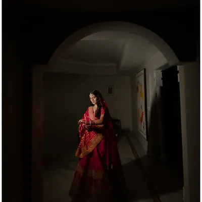 A dramatic bridal portrait using shadow and light. The bride, in her radiant red lehenga, is framed by an archway, creating a sense of mystery and timeless beauty.