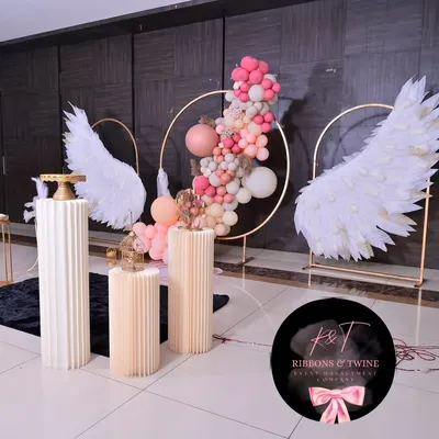 A beautiful baptism setup featuring angel wings, a central gold ring with a balloon garland, and fluted plinths for the cake and other displays. The color scheme of pink, peach, and white creates a soft and celebratory feel.