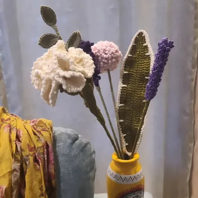 A beautiful, long-lasting crochet flower arrangement in a bright yellow vase. This handmade bouquet includes a detailed white carnation, a fluffy pink pom-pom, a sprig of purple lavender, and textured green leaves, offering a cheerful and permanent decorative accent.