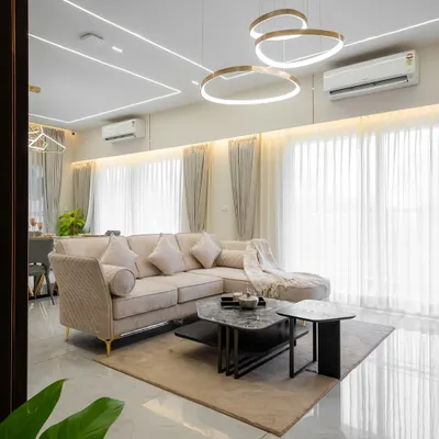 Natural light floods this living room, highlighting the clean lines of the ceiling and the elegant circular chandeliers. The neutral-toned sectional sofa and layered rugs create a calm, inviting atmosphere perfect for relaxation and quiet conversations.