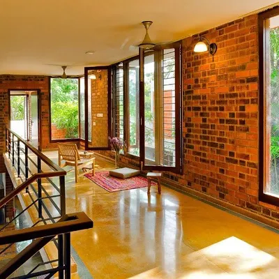 The upper-level corridor at the Vidyut Kapoor Residence features a continuous exposed brick wall, complemented by large windows that offer views of the surrounding greenery. The polished yellow oxide flooring reflects the warm tones and natural light.