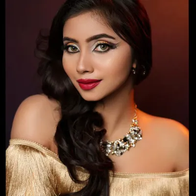 A glamorous studio portrait with dramatic lighting. The focus is on the model's striking makeup, statement necklace, and the texture of the gold off-shoulder top.