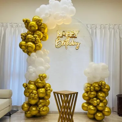An elegant birthday decoration in gold and white. This setup features an organic balloon arch, a "Happy Birthday" neon sign against a white panel, and a stylish pedestal for the cake.