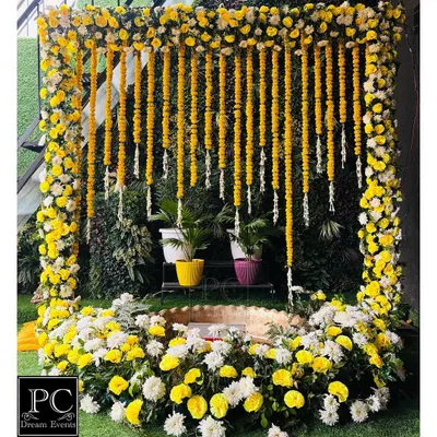 A classic Haldi setup featuring a frame of yellow and white flowers with hanging marigold garlands. The central seating area is designed for the ceremony, surrounded by lush greenery for a fresh, natural feel.