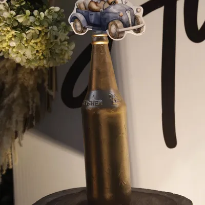 A creative decor detail for a teddy bear themed party. A small cutout of a teddy bear in a vintage car is placed atop a painted bottle, sitting on a rustic wood slice.