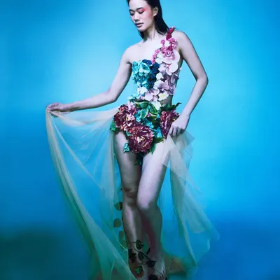 A full-length portrait of Ophelia against a solid blue background. The custom garment, adorned with flowers, is clearly visible, showcasing the intricate styling and the model's graceful pose.