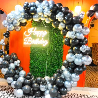 This is a great example of a sophisticated party backdrop for adults. A full ring of black and silver chrome balloons frames a green foliage wall, with a neon "Happy Birthday" sign in the center, creating a perfect spot for selfies and group photos.