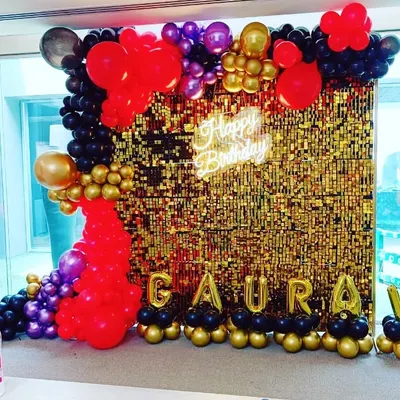 A vibrant and glamorous birthday backdrop. This setup features a gold shimmer wall framed by a dramatic balloon garland in red, black, purple, and gold, with a 'Happy Birthday' neon sign.