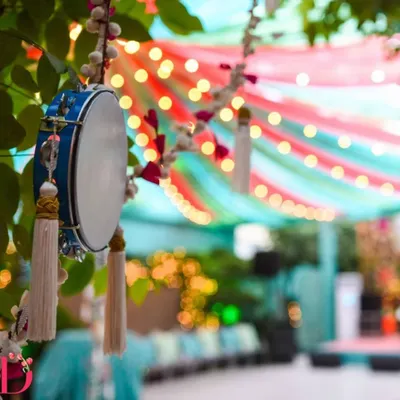 A beautiful detail from a festive decor setup, featuring a traditional dafli (tambourine) with tassels hanging from a tree. The background shows colorful drapes and string lights, creating a vibrant ambiance.
