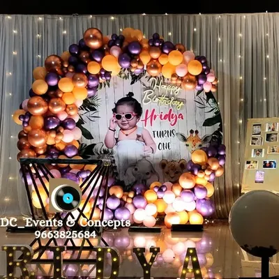 A stylish first birthday setup with a circular backdrop featuring the baby's photo, a modern balloon arch in metallic tones, and illuminated marquee letters spelling out the name.