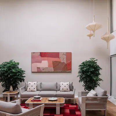 This living area in the Cocoon House is defined by its soaring double-height ceiling and a calm, neutral palette. We introduced warmth and personality with a large abstract painting in soft pinks, woven-texture furniture, and lush indoor plants, creating a space that feels both expansive and inviting.