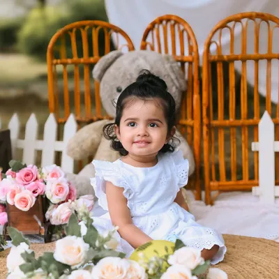 A close-up of a sweet baby girl during her first birthday photoshoot. Her gentle smile is captured perfectly in this outdoor setup, surrounded by soft pink flowers and a giant teddy bear.