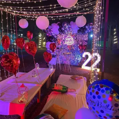 A beautiful outdoor cabana setup for a 22nd birthday. The space is decorated with fairy lights, heart-shaped foil balloons, and a statement backdrop with a neon sign and marquee numbers.