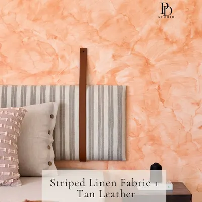 This bedroom detail shows a beautiful combination of textures: a striped linen headboard with a tan leather strap, set against a warm, textured paint wall. This pairing creates an earthy, tactile, and inviting atmosphere.
