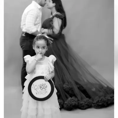 A creative family portrait where the older sister holds a sign announcing her promotion to "BIG Sister," while her parents share a kiss in the background.