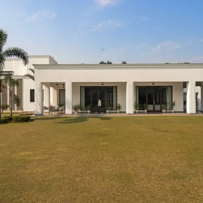 An exterior view of the Mehalin Farmhouse, where the landscape design emphasizes a vast, open lawn to accentuate the building's neo-classical form. The placement of palm trees frames the structure without obstructing the view.
