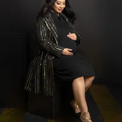 A touch of gold adds instant glamour. Here, the mom-to-be is seated, looking down at her bump, wearing a chic black dress paired with a sparkling gold-striped jacket for a sophisticated look.
