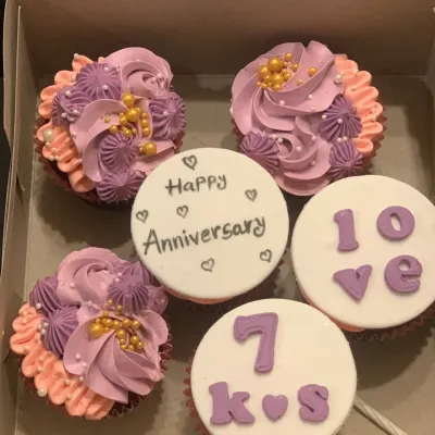 A custom anniversary cupcake box. This set includes intricately piped floral cupcakes in lavender and pink, alongside fondant-topped cupcakes that spell out a personal message of love for a 10th anniversary.