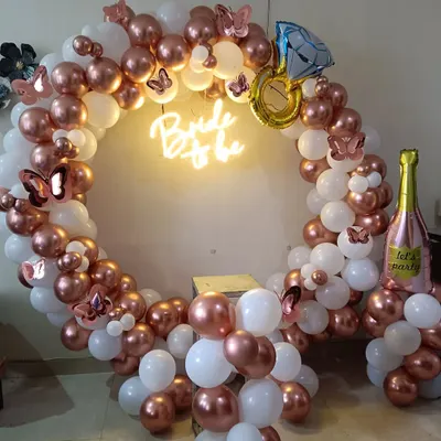 This is a complete bridal shower photo booth setup. It includes a balloon ring in rose gold and white, a "Bride to be" neon sign, and fun props like butterfly cutouts and foil balloons shaped like a diamond ring and a champagne bottle.