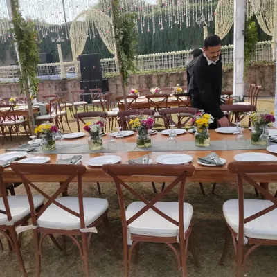 Long wooden tables paired with elegant cross-back chairs and padded cushions, creating a rustic-chic dining experience for an outdoor event.