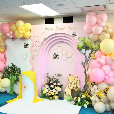 A sweet and charming Winnie the Pooh-themed setup for an Aqeeqah celebration, featuring a pink arched backdrop with honeycomb details. The scene includes a Pooh Bear cutout, a "hunny" tree, and a lovely balloon garland in pastel pink, yellow, and cream.