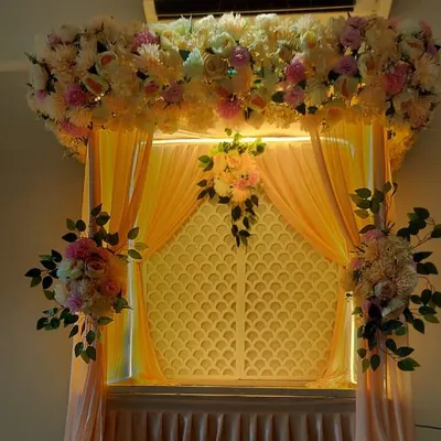 This decoration uses a warm, peachy color scheme with soft drapes and a thick arrangement of pastel-colored flowers. The integrated lighting from above and behind the lattice panel gives the entire setup a gentle, warm glow.