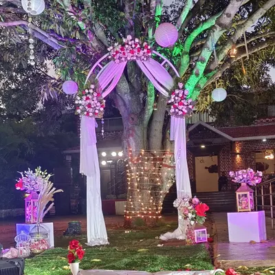 I love transforming outdoor spaces, like this garden proposal setup under a large tree. The design includes a floral arch draped with soft fabric, hanging lanterns, and a rose petal aisle, using colored lights to create a magical atmosphere after sunset.