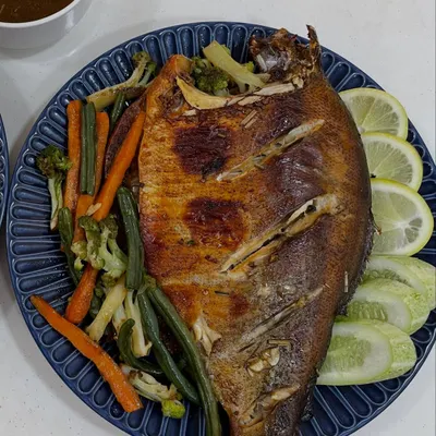 A whole roasted fish, marinated in our house Naga spices and roasted until the skin is crisp. It's served on a beautiful blue plate with steamed vegetables, fresh lemon, and a dipping sauce.