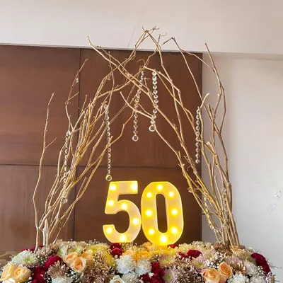 A four-foot-long floral centerpiece I created for a 50th birthday. It features light-up marquee numbers, an arch of gold-painted branches with hanging crystals, and a bed of roses and chrysanthemums.