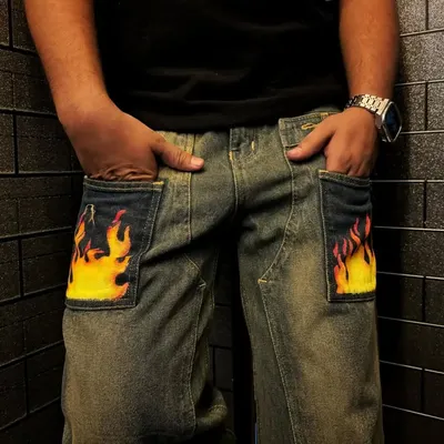 A pair of baggy denim jeans featuring hand-painted flames on the front pockets. The bright yellow and orange design adds a classic hot-rod feel to the pants.