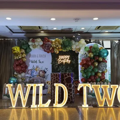 A full view of the "Wild Two" jungle safari stage we created for twins Vihaan and Vaniya. The setup includes a central balloon arch, a green foliage backdrop with a neon sign, and character cutouts, all framed by our popular light-up marquee letters.