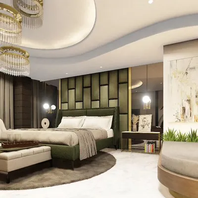 A luxurious master bedroom in a Noida penthouse, featuring a custom olive green upholstered headboard with geometric paneling. The room is designed with a curved false ceiling, elegant chandeliers, and a dedicated seating area for a suite-like feel.