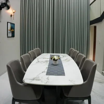 This dining setup is designed for grand entertaining. The large, marble-finish tabletop provides a luxurious surface, surrounded by ten fully upholstered chairs for maximum comfort. The floor-to-ceiling curtains add a touch of drama and sophistication to the double-height space.